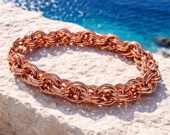May include: A copper-colored bracelet with a chainmail design. The bracelet is made of interlocking circular links, creating a textured and detailed appearance. The jewelry is displayed on a light-colored stone surface with a blue ocean background.