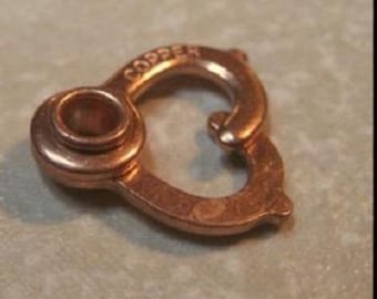 May include: A heart-shaped copper jewelry clasp featuring a circular eyelet on one side and a spring-loaded gate mechanism on the other. The word COPPER is embossed in capital letters along the top curve of the metallic fastener.
