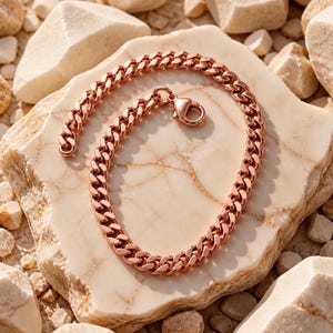 May include: A rose gold-toned curb chain bracelet featuring interlocking flat, polished links. The bracelet is circular in shape and includes a secure lobster claw clasp mechanism at one end, showcasing a metallic finish with warm copper hues.