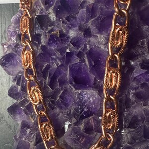 May include: A copper-colored chain necklace with oval links, each featuring an embossed design. The necklace is displayed against a backdrop of a large, textured, purple amethyst crystal, creating a contrast of colors and textures.