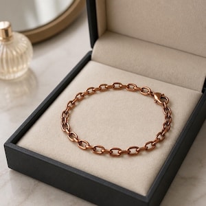 May include: A copper-colored chain bracelet with an oval link design rests inside a black jewelry box with a cream-colored interior. The bracelet is secured with a lobster clasp. A perfume bottle is visible in the background.