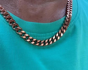 May include: A rose gold chain necklace with a curb link design. The necklace is displayed against a teal green shirt, highlighting the warm tones of the jewelry. The chain is thick and appears to be made of metal.