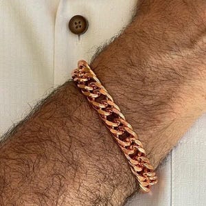 May include: A copper-colored chain link bracelet. The bracelet is made of thick, interlocking links. The bracelet is worn on a wrist, and the background includes a white shirt.