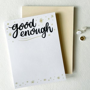 May include: White card with gold stars and the text "good enough" in black cursive lettering. The card is on top of a beige card.