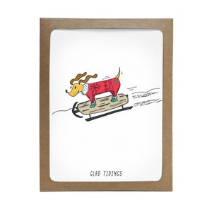May include: A Christmas card featuring a cartoon dog wearing a red sweater with a snowflake pattern, riding a sled down a snowy hill. The card has the text "Glad Tidings" printed on the bottom.