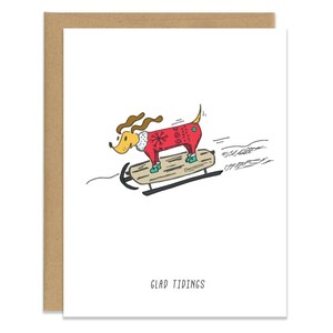 May include: A cartoon illustration of a dog wearing a red sweater with a snowflake pattern, riding a sled with the text "Glad Tidings" below.