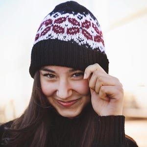 May include: A black knit beanie with a geometric pattern in white and burgundy. The beanie is worn by a person wearing a black sweater. The person is smiling and touching their face.