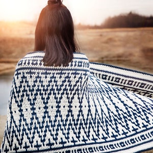 May include: A white and navy blue crocheted blanket with a geometric pattern. The blanket is draped over a person's shoulders, with the person's back to the camera. The background is a blurred outdoor scene.