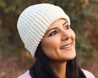 May include: A woman wearing a white ribbed knit beanie and a light pink cardigan sweater. The sweater has a V-neck and button closure. The background is a blurred view of trees and tall grass.