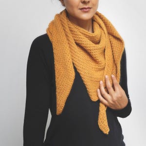 Può includere: Sciarpa lavorata a maglia giallo senape a forma triangolare. La sciarpa è drappeggiata sulle spalle e sul petto. La trama a maglia è visibile e la sciarpa è realizzata in un materiale morbido e caldo. La persona indossa una maglietta nera a maniche lunghe.