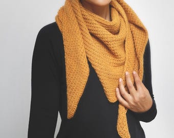 May include: Mustard yellow knitted shawl with a triangular shape. The shawl is draped over the shoulders and chest. The knitted texture is visible, and the shawl is made of a soft, warm material. The person is wearing a black long-sleeve shirt.
