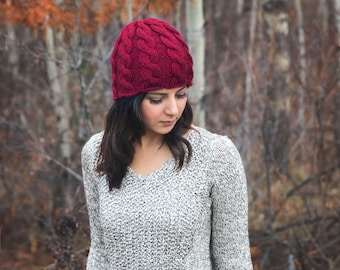 May include: A woman wearing a red knitted beanie with a cable knit pattern. She is wearing a gray sweater and is standing in front of a blurred background of trees.