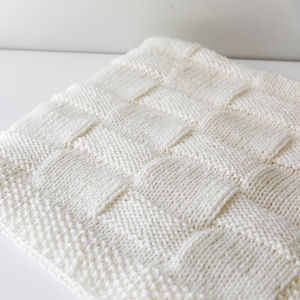 May include: Cream-coloured knitted blanket with a basket weave design. The blanket is made of soft yarn and has a textured surface. The blanket is folded and displayed on a white surface.