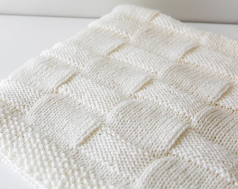 May include: Off-white knitted blanket with a woven basket weave pattern. The blanket is made of soft yarn and has a textured surface. The blanket is folded and displayed on a white surface.