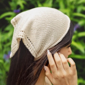 May include: A cream-coloured knit headscarf featuring a triangular shape with a textured stockinette stitch pattern and a decorative border along the edges. The fabric is made of a soft yarn, creating a lightweight and flexible accessory for the head.