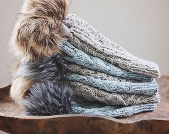 May include: A stack of knitted hats with faux fur pom-poms. The hats are in shades of grey and blue, with a cable knit design. The pom-poms are brown and grey. The hats are stacked on a wooden stool.