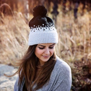 May include: A woman wearing a gray knitted hat with a black pom pom and a white and black heart pattern. The hat is knit with a gradient of white to black.