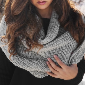 May include: A light gray, chunky knit infinity scarf. The scarf is made of a thick yarn and has a textured appearance. The scarf is wrapped around the neck. The person is wearing a black top.
