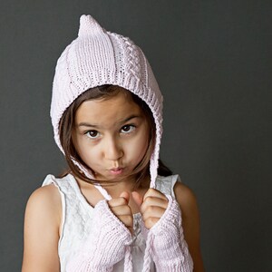 May include: A young girl wearing a pink knitted hat with a pointed top and matching fingerless gloves. The hat has a cable knit pattern and the gloves have a simple knit pattern.
