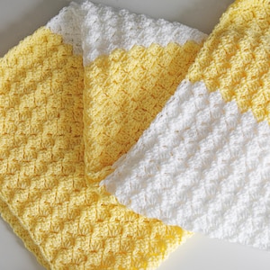 May include: Close-up of three handmade crochet blankets. Two blankets are yellow, and one is white. The blankets are folded and layered, showcasing the textured stitch pattern. The blankets are soft and cozy.