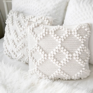 May include: Two white crocheted throw cushions with a textured diamond pattern and raised bobbles. The cushions are on a white faux fur surface.