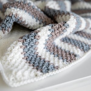 May include: A close-up of a handmade crochet blanket. The blanket features a repeating pattern of horizontal stripes in white, light blue, and brown. The texture of the crochet stitches is visible, creating a soft and cosy appearance. The blanket is folded, showing its thickness and construction.