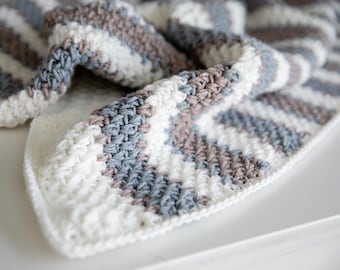 May include: A close-up of a handmade crochet blanket. The blanket features a repeating pattern of horizontal stripes in white, light blue, and brown. The texture of the crochet stitches is visible, creating a soft and cozy appearance. The blanket is folded, showing its thickness and construction.