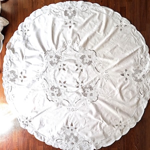 May include: A white, round tablecloth with intricate floral and geometric cutwork and embroidery. The scalloped edge adds a decorative touch. The tablecloth is made of a soft, white fabric and is suitable for a dining table.