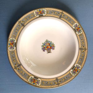 May include: A round white ceramic bowl featuring a central colorful fruit basket illustration. The wide rim is decorated with a repeating pattern of blue and yellow scrollwork, interspersed with small fruit basket motifs and a gold-toned edge.