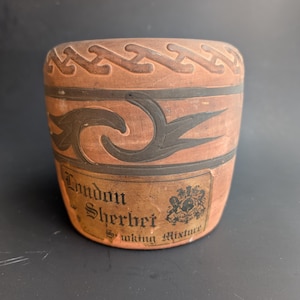 May include: A cylindrical terracotta tobacco jar featuring a dark brown embossed geometric pattern around the rim and a stylized leaf design in the center. A rectangular label reads London Sherbet Smoking Mixture with a crest illustration.