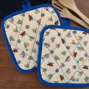 May include: Two blue and white quilted pot holders with a pattern of small green and brown characters. The pot holders are on a wooden surface.