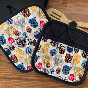 May include: Two black and white quilted pot holders with a pattern of colorful Mandalorian helmets. The pot holders have a black trim and a loop for hanging.