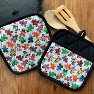 May include: Two black and white quilted pot holders with a superhero gingerbread man pattern. The pot holders have a loop for hanging.