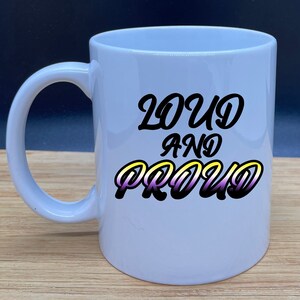 Puede incluir: Taza de cerámica blanca con un gráfico negro que dice "Loud and Proud" en una fuente estilizada. La palabra "Proud" está en un degradado de amarillo, morado y negro.