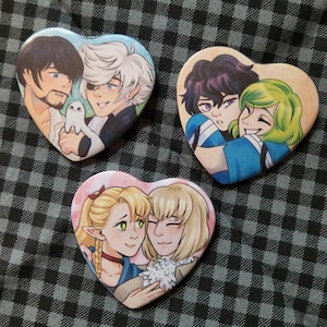 May include: Three heart-shaped buttons featuring colorful illustrations of anime-style characters. Each button showcases a different pair of characters in a loving embrace, with varying skin tones, hair colors, and clothing styles. The background is a checkered pattern.