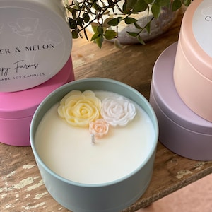 May include: A teal candle jar with a white soy candle and three decorative flower accents. The flowers are yellow, white, and orange. The candle jar has a label that reads "Happy Farms" and "Water & Melon" with the text "Jade Soy Candles".
