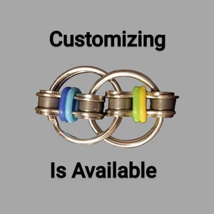 Peut inclure: Un jouet de fidget argenté avec des accents bleus et verts. Le jouet est composé d'anneaux et de composants cylindriques imbriqués. Le texte "Customizing Is Available" est affiché en noir.