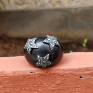 Puede incluir: Un anillo negro y redondeado con tres adornos de estrellas plateadas. Las estrellas están incrustadas con pequeñas piedras brillantes. El anillo está sobre un fondo borroso.