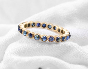May include: A gold band ring with a row of blue gemstones. The ring is on a white fabric background.