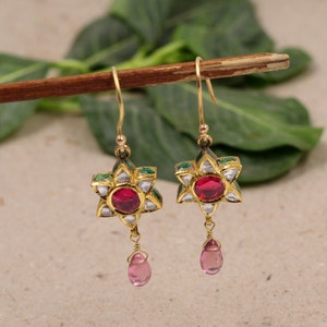 May include: A pair of gold earrings with a star-shaped design. The earrings are adorned with red and white gemstones and green enamel. Each earring has a pink teardrop dangling from the bottom.