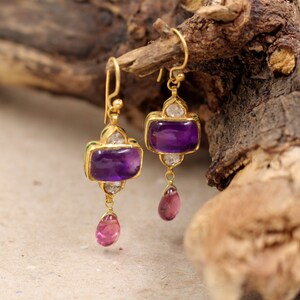 May include: A pair of gold earrings with amethyst and pink stones. The earrings have a teardrop shape with a small amethyst stone at the top and a larger amethyst stone in the middle. The earrings are accented with small diamonds.