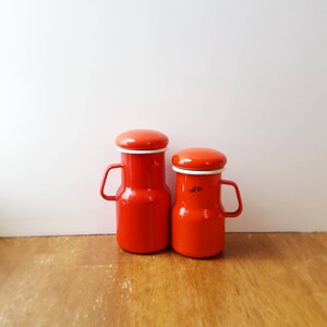 May include: Two orange enamelware canisters with white lids and handles. The canisters have a vintage look and are perfect for storing spices or other kitchen essentials.