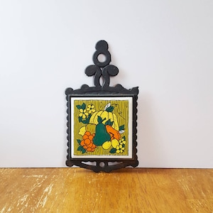 May include: Black cast iron trivet with a colorful ceramic tile insert featuring a still life of fruits and flowers. The tile shows a yellow pumpkin, green pear, orange fruit, and yellow flowers. The trivet has a decorative handle.