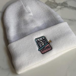 May include: White beanie with a small embroidered design of a can of sardines. The can is pink and blue with the word "SARDINES" printed on it.