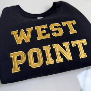 Puede incluir: Sudadera negra de cuello redondo con las palabras "WEST POINT" bordadas en letras doradas. La sudadera está doblada y parece estar hecha de un material suave. Las letras doradas tienen un estilo universitario texturizado.