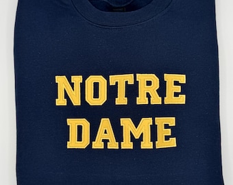 Może przedstawiać: Granatowa bluza z napisem "NOTRE DAME" w złotych literach. Ubranie jest starannie złożone, podkreślając tekst. Klasyczny design, idealny dla fanów uniwersytetu.