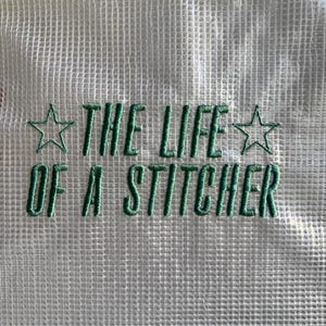 Puede incluir: Primer plano de una tela de malla blanca con texto bordado en verde y detalles de estrellas. El texto dice "THE LIFE OF A STITCHER", lo que sugiere un tema de artesanía o afición. El bordado es detallado y la estética general es limpia.