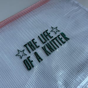 May include: A clear mesh zipper pouch with the text "THE LIFE OF A KNITTER" embroidered in green. The pouch has a red-orange trim and a small white tag.