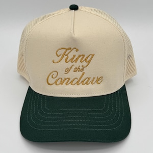 Op de afbeelding: Een beige en donkergroene truckerpet met de gouden tekst "King of the Conclave". De pet heeft een mesh achterkant en een gebogen klep. De bovenkant is beige en de klep is donkergroen.