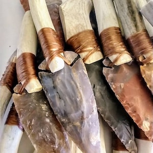 May include: A collection of handmade flint arrowheads with antler handles wrapped in brown leather cord. The arrowheads are various shades of brown, gray, and white.